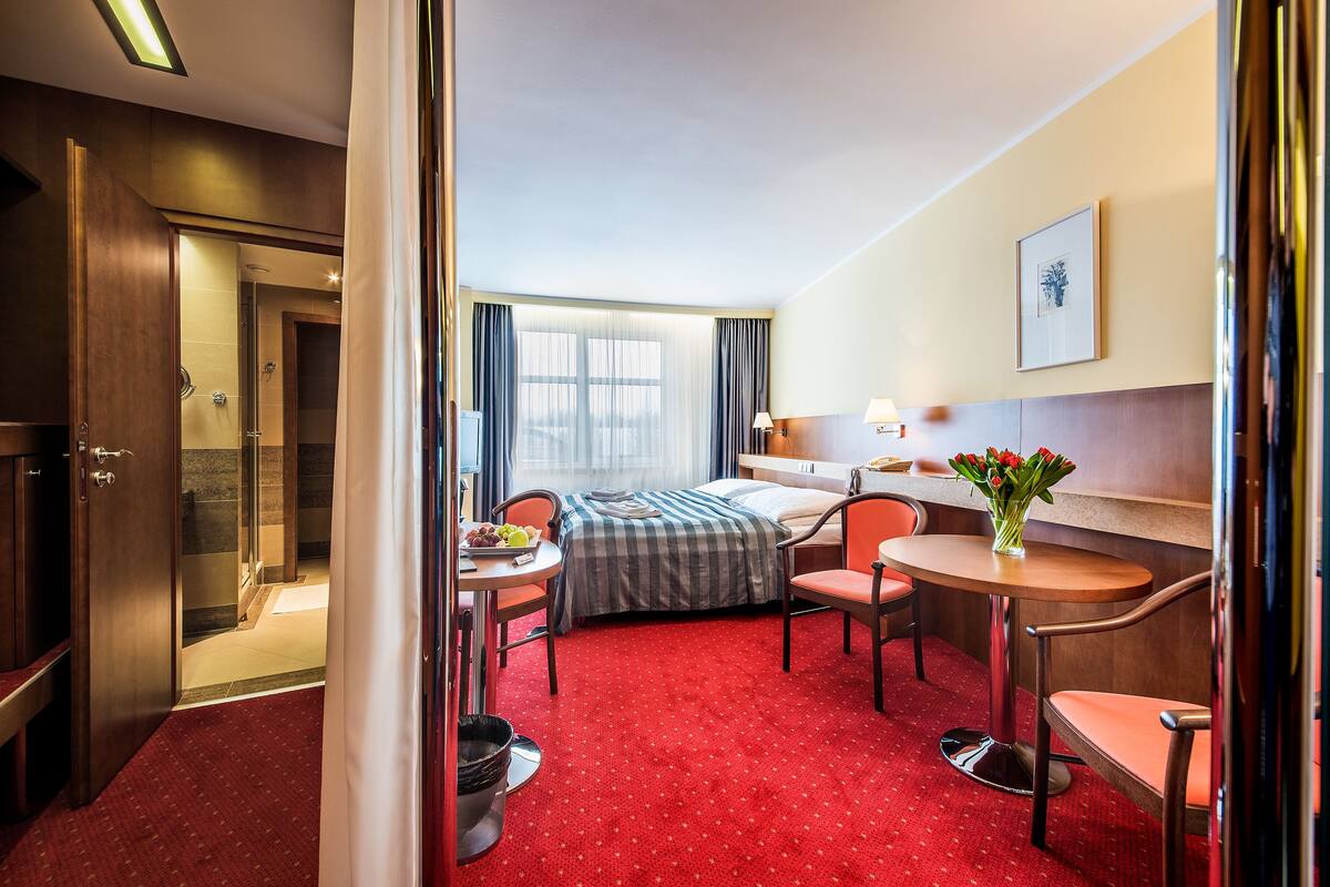 A hotel room with a bed, a desk, a chair, a small table, a vase with flowers, and a shower area.