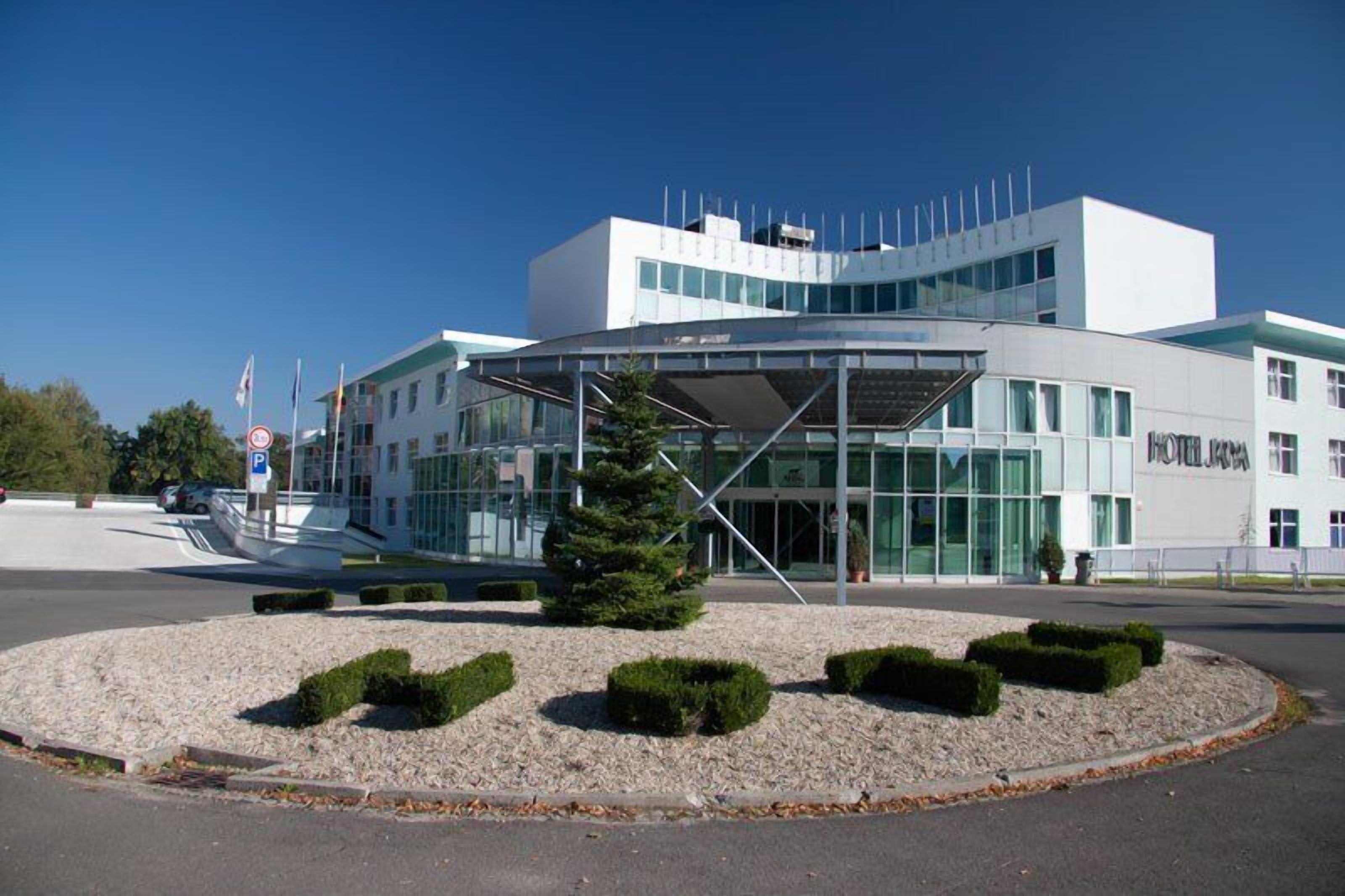 Et moderne hotel med glasfacade, overdækket indgang og anlagt område med buske og et lille træ.