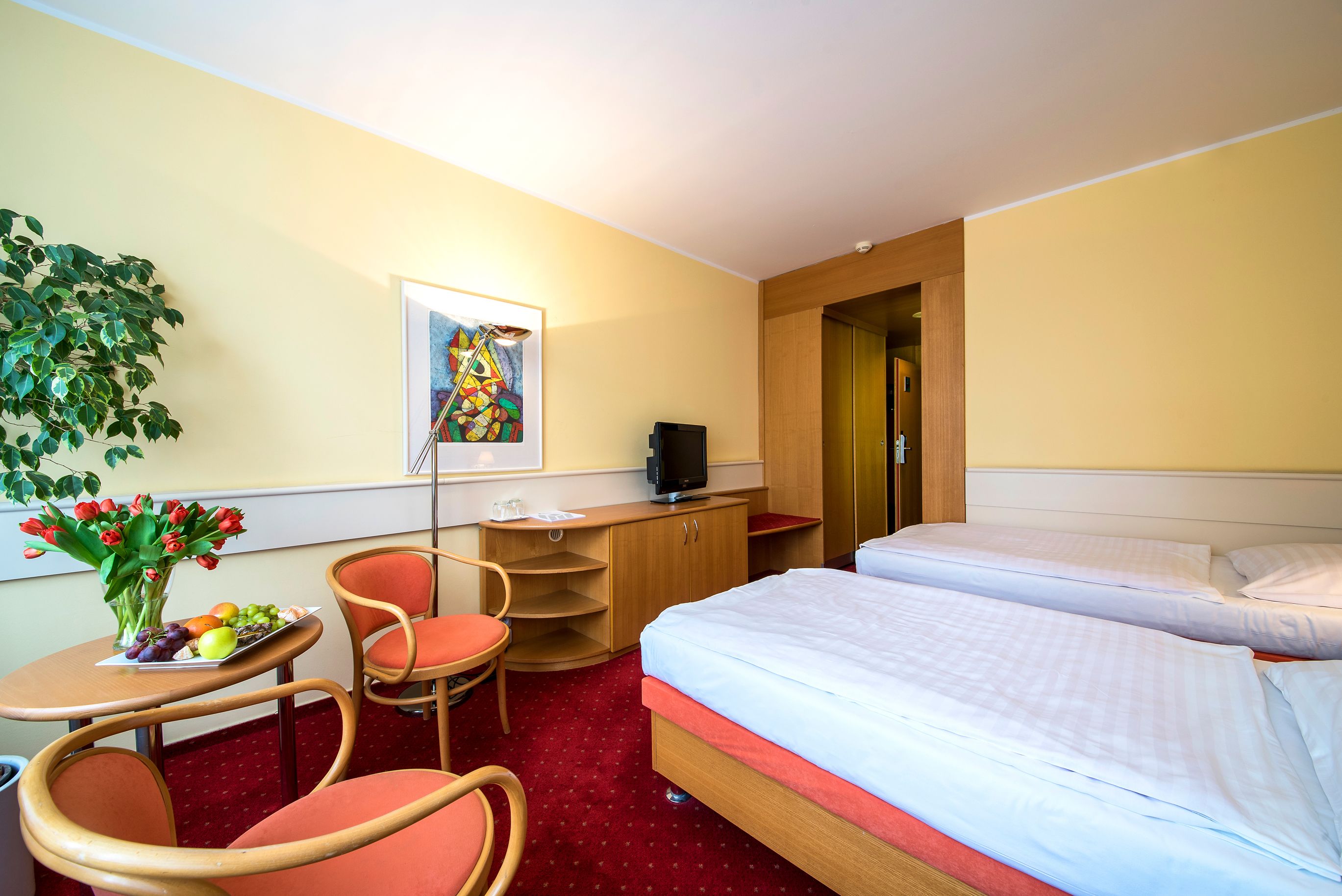 A hotel room with two beds, a desk with a chair, a television, a plant, and a painting on the wall.