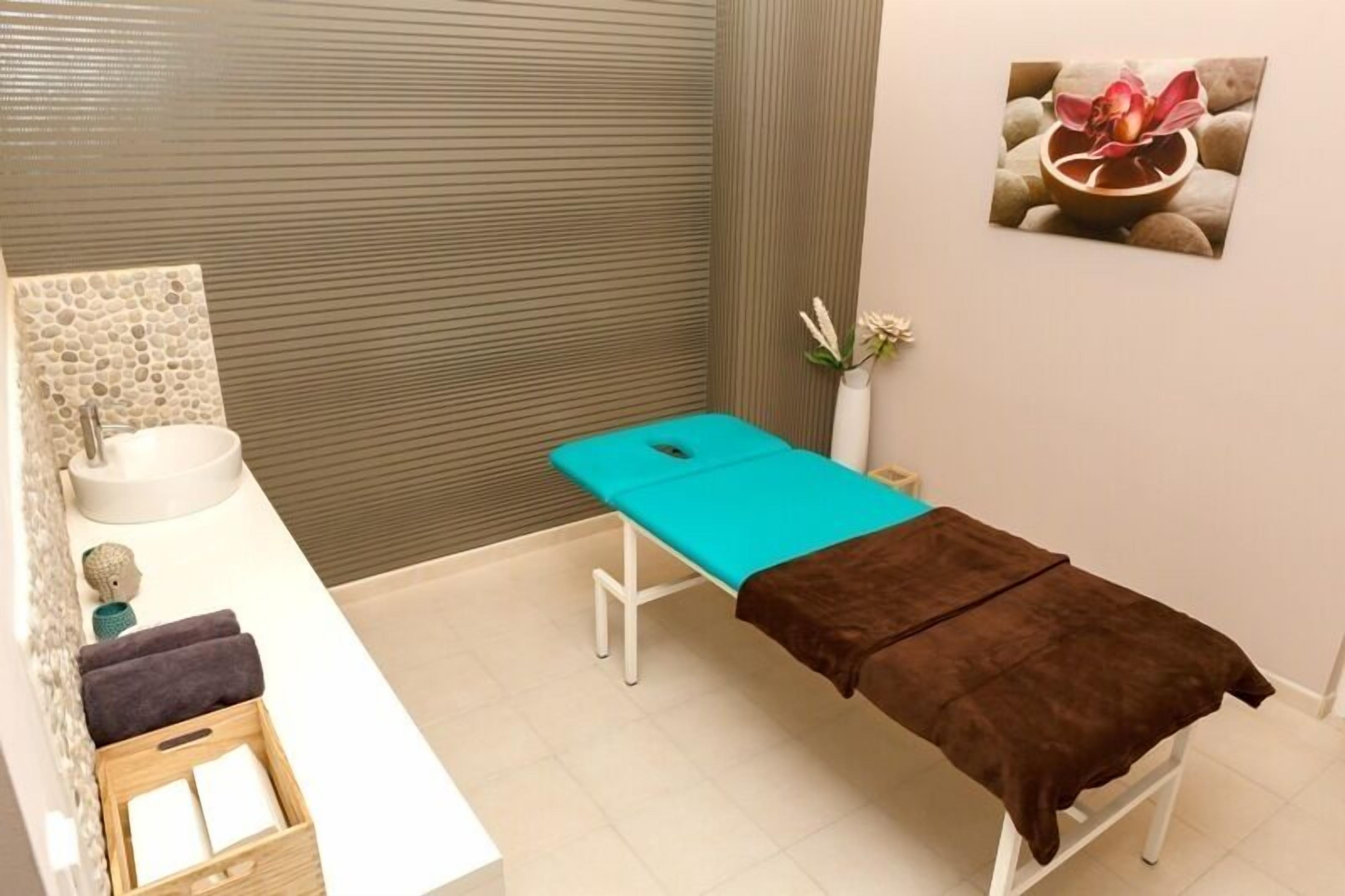 Een spa ruimte met een massagetafel, een wastafel, een klein houten doosje met handdoeken en een decoratieve vaas met bloemen.