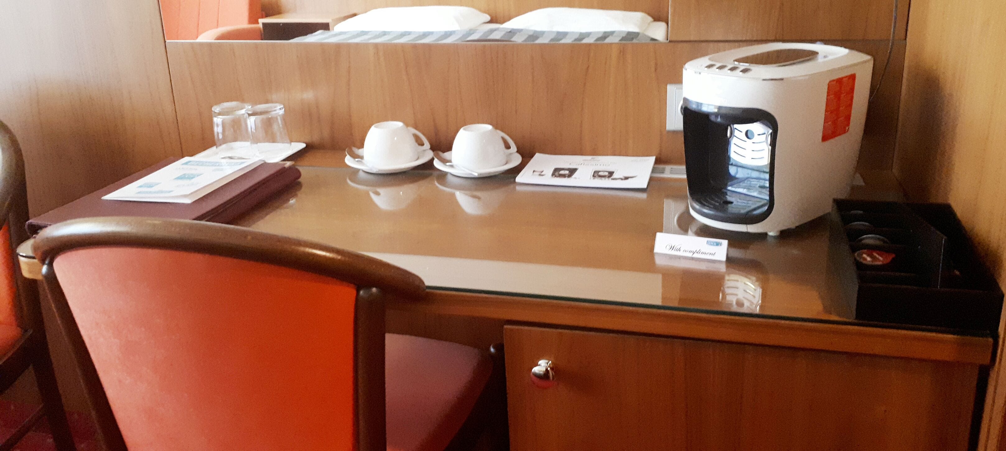 A hotel room with a wooden desk, a coffee maker, two white cups, a glass, and a red chair.