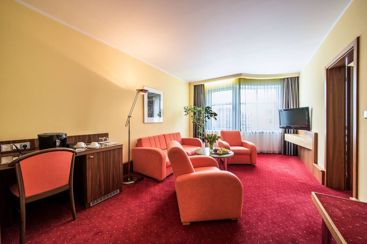 A hotel room with a red carpet, a sofa, a chair, a desk with a coffee maker, a TV, and a window with curtains.