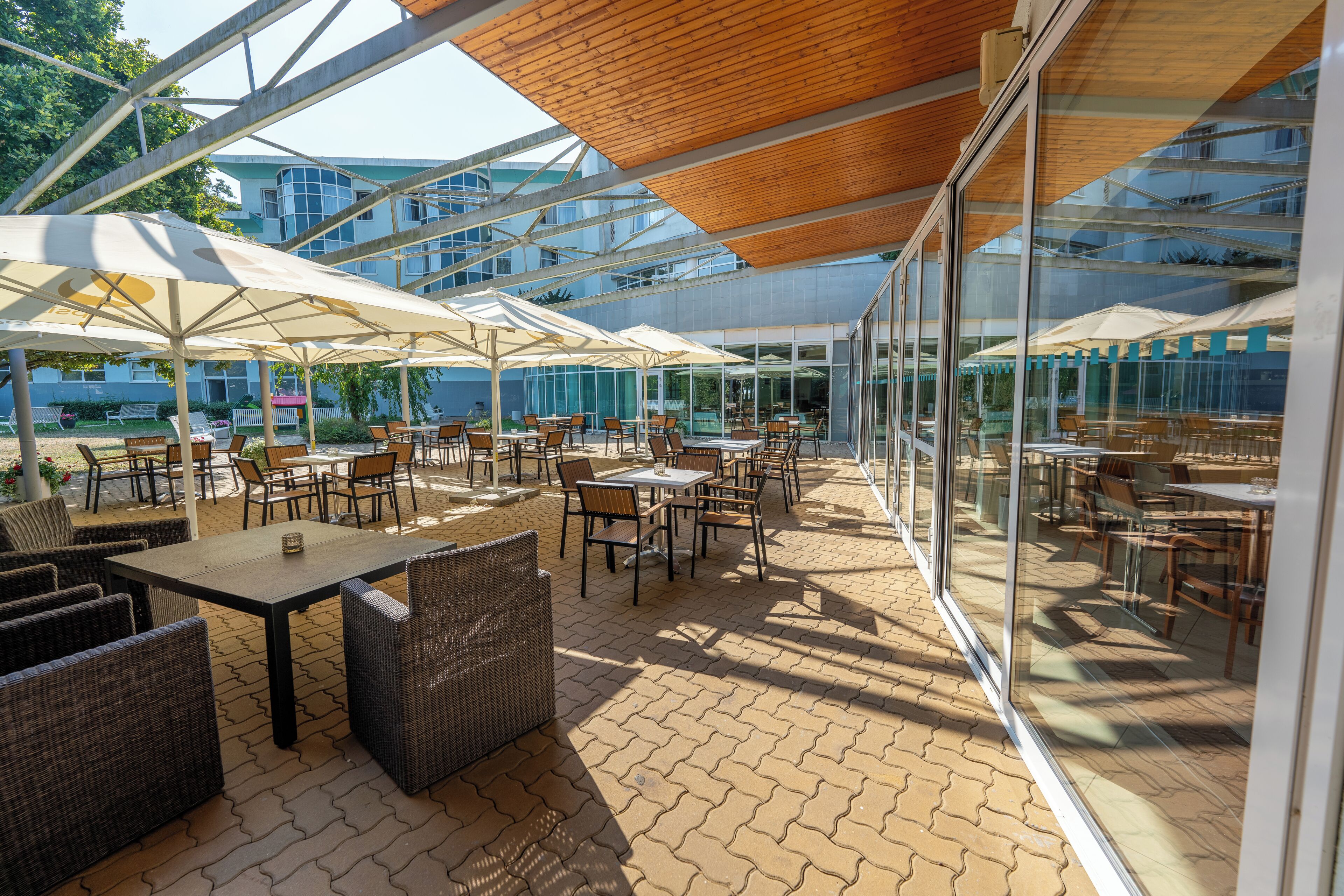 An outdoor patio area with tables and chairs, covered by umbrellas, and large windows offering views of the surrounding buildings.