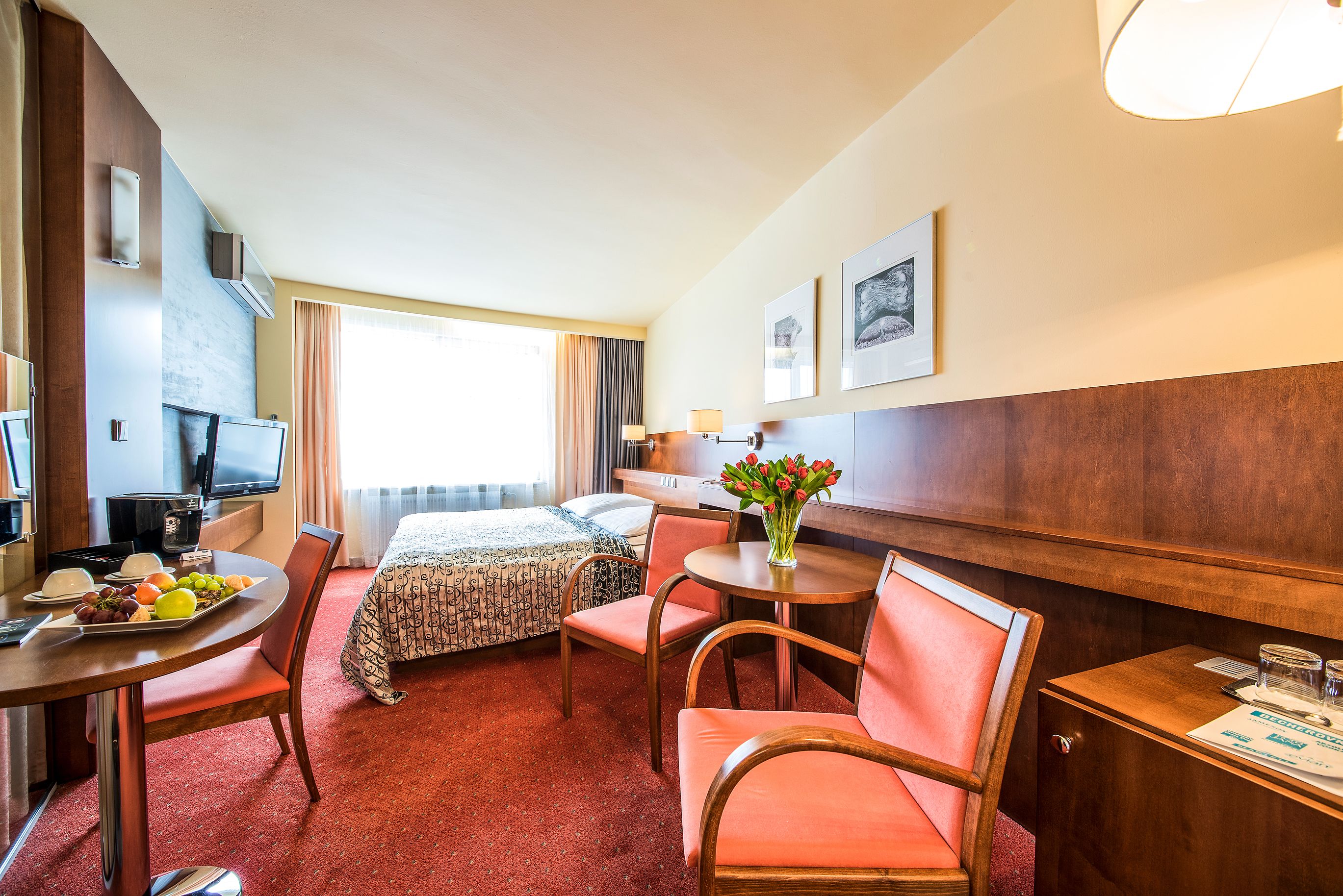 A hotel room with a bed, a desk with a chair, a dining table with a chair, a TV, and a vase with flowers.