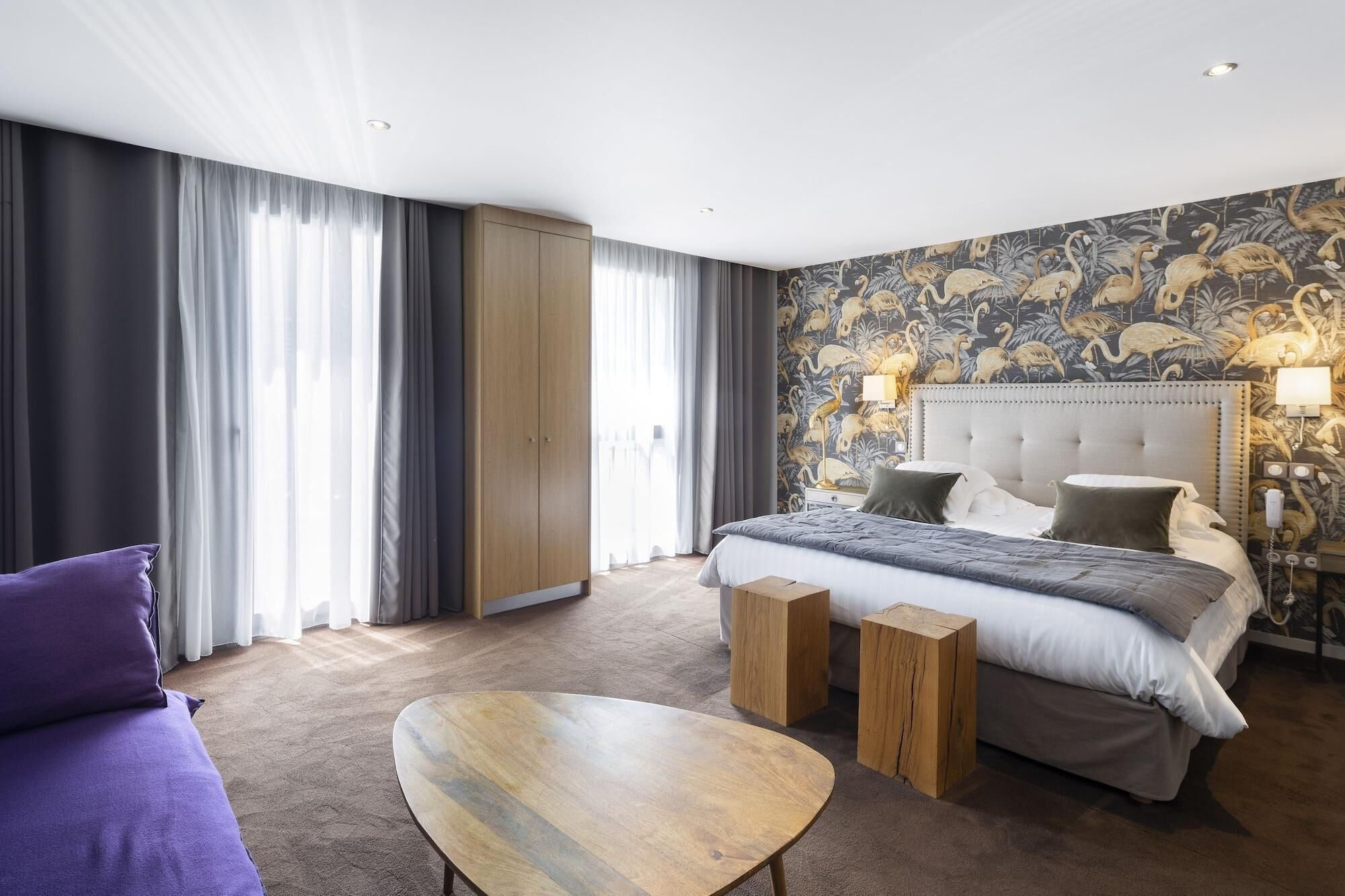 A modern hotel room with a large bed, a wooden coffee table, and a view of the city through floor-to-ceiling windows.
