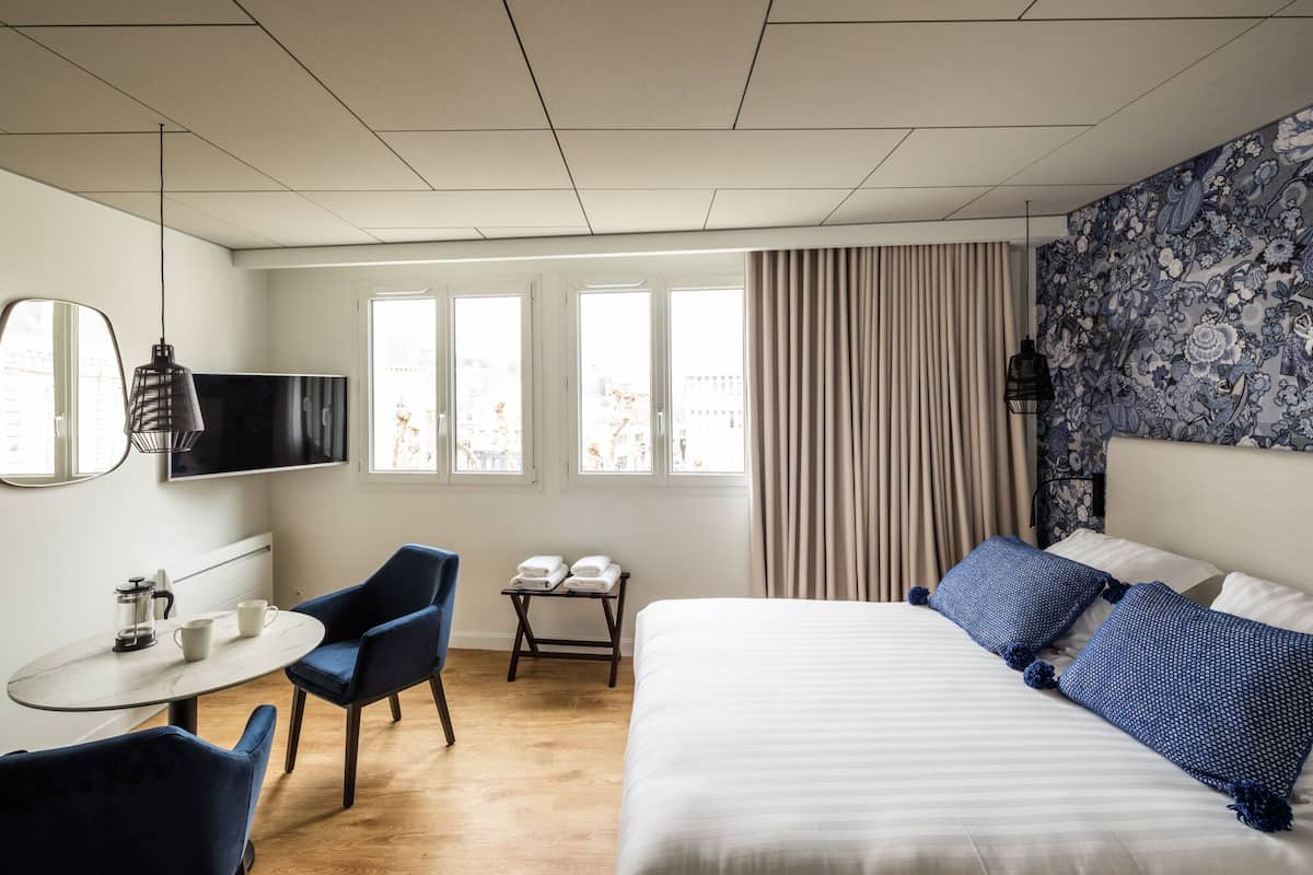 A hotel room with a bed, a desk, a chair, a TV, and a window with curtains.