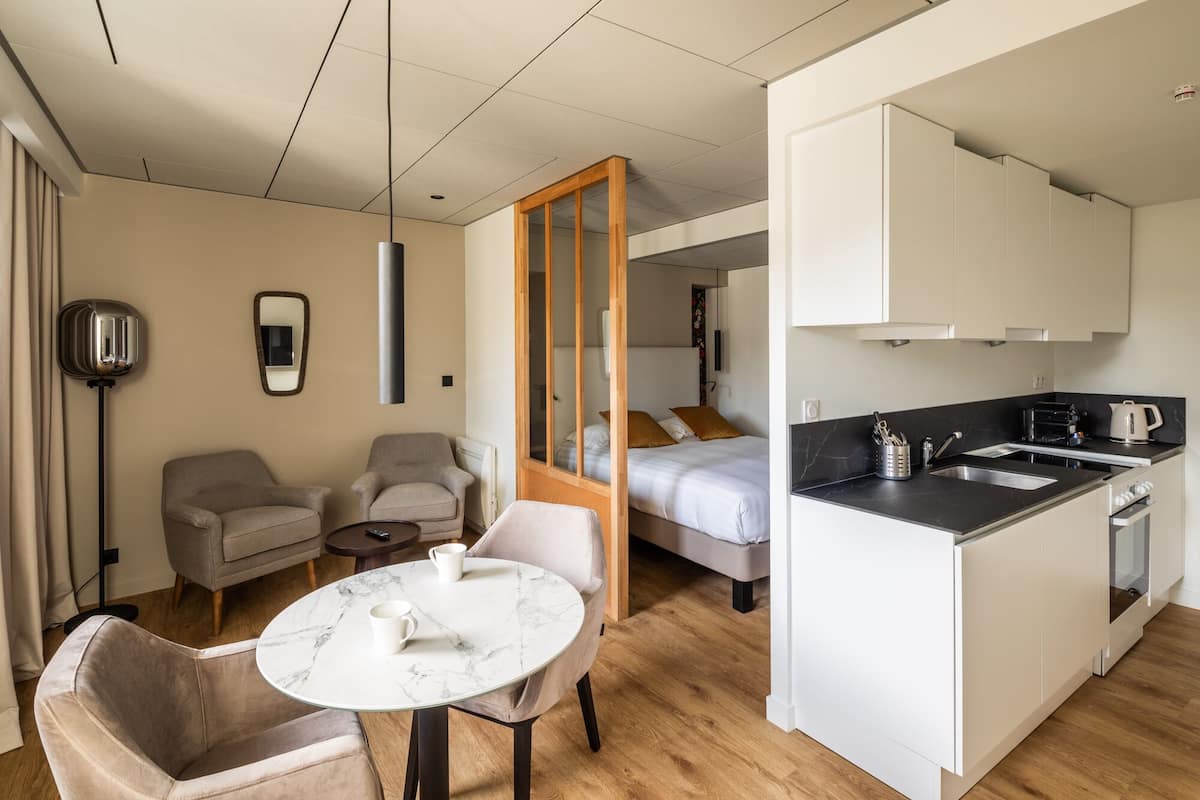 A compact hotel room with a bed, a seating area with two chairs and a table, a kitchenette, and a large mirror.
