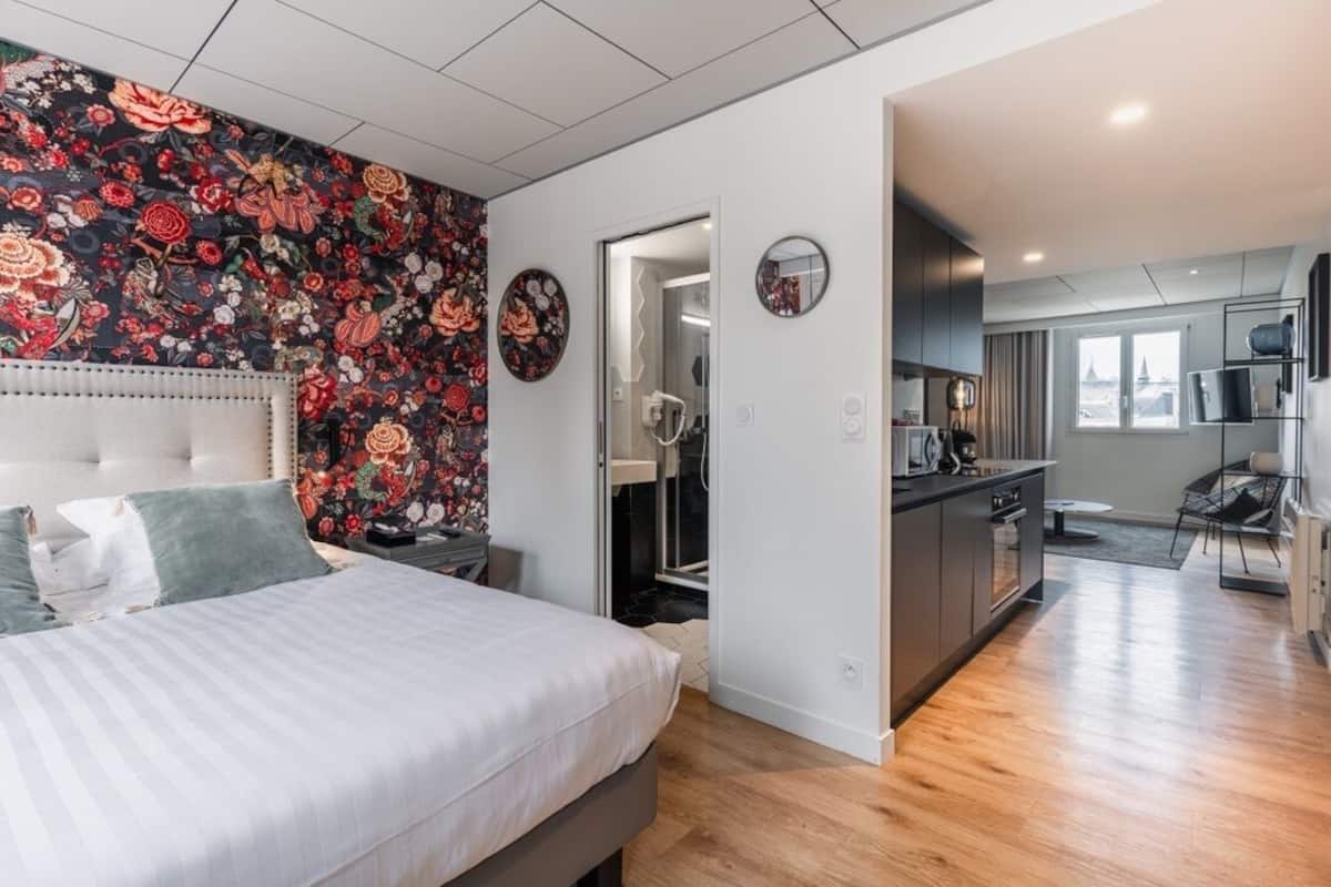 A modern hotel room with a large bed, a floral-patterned wall, a kitchen area, and a view of the city.