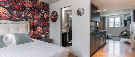 A modern hotel room with a large bed, a floral-patterned wall, a kitchen area, and a view of the city.