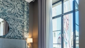 Et hotelværelse med en stor seng, en sengegavl med et spejl, et vindue med udsigt til bygninger og en væg med et bladmotiv.