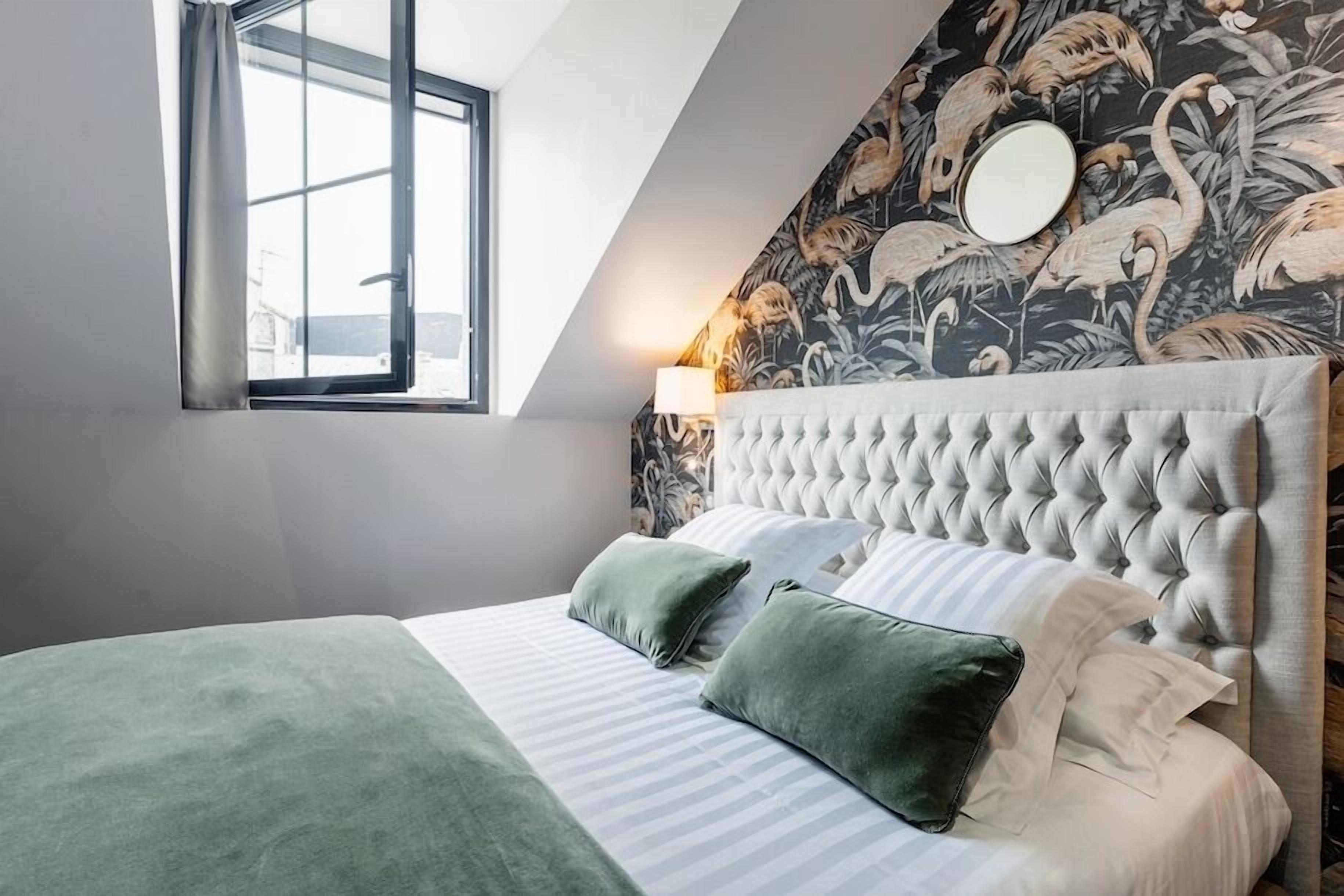 Une chambre à coucher avec une tête de lit capitonnée, une grande fenêtre et un mur recouvert d’un papier peint aux motifs tropicaux.