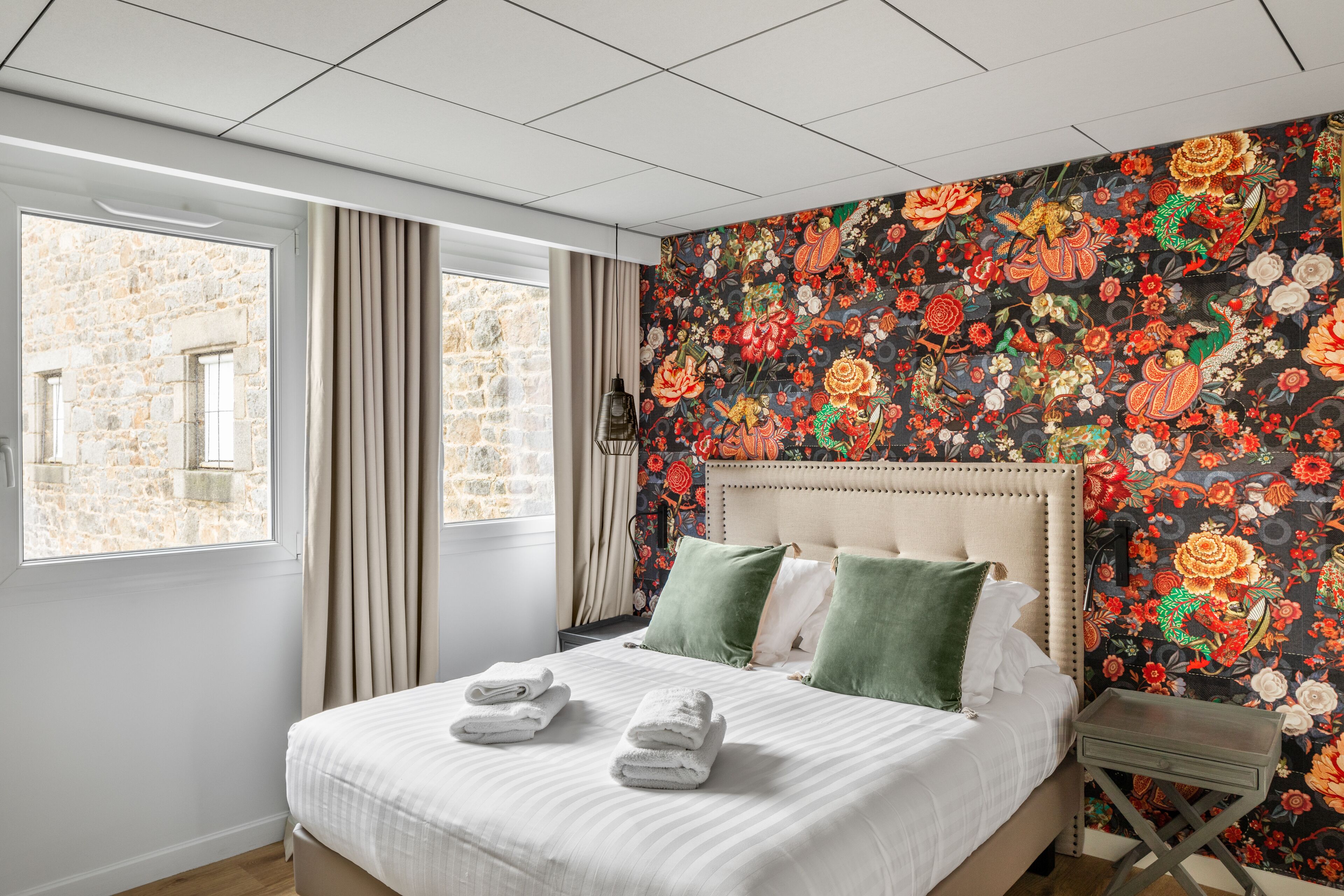 A hotel room with a large bed, a view of stone buildings through the window, and a floral wallpaper.