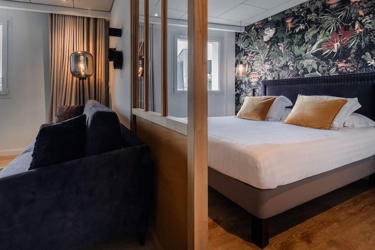 A hotel room with a bed, a sofa, a mirror, and a wall with a floral pattern.
