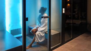 A person in a white bathrobe sitting on a bench in a glass-walled room with a blue light.