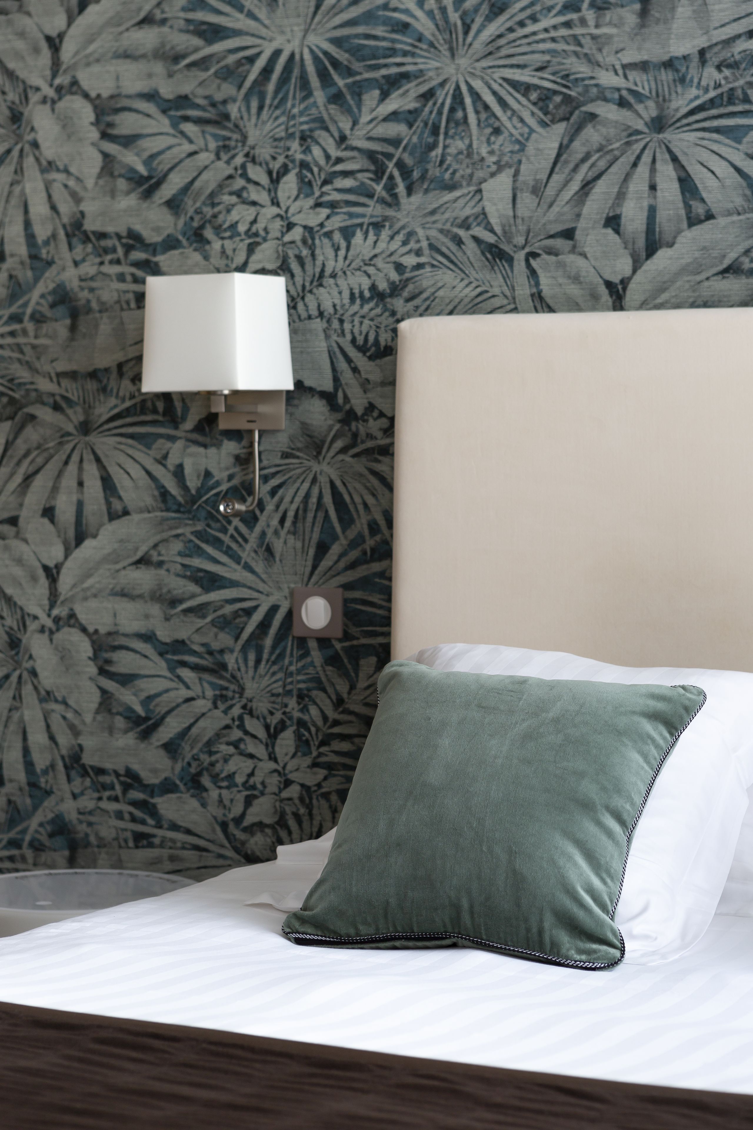 A bed with a white mattress and a green pillow, a wall-mounted lamp, and a floral wallpaper pattern.