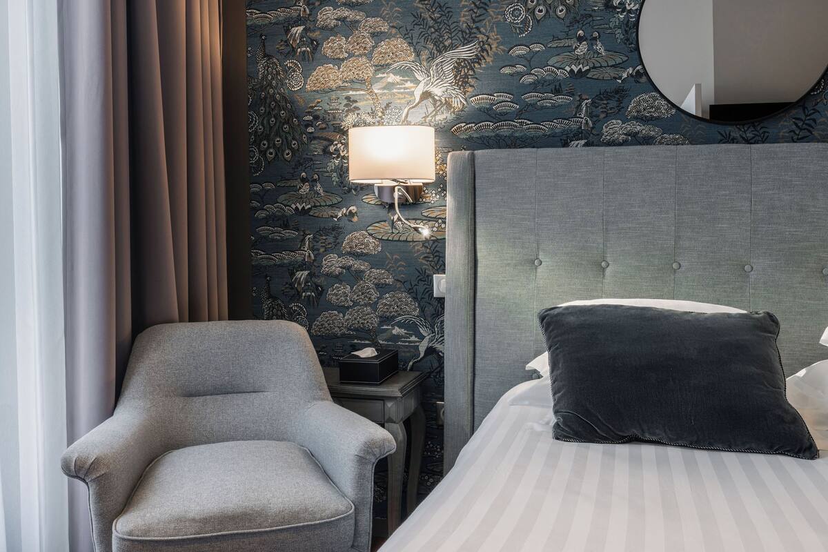 A hotel room with a bed, a chair, a nightstand, a lamp, and a wall with a detailed mural.