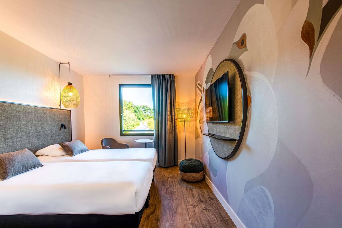 A hotel room with two beds, a TV, a window with curtains, a small table, and a lamp.