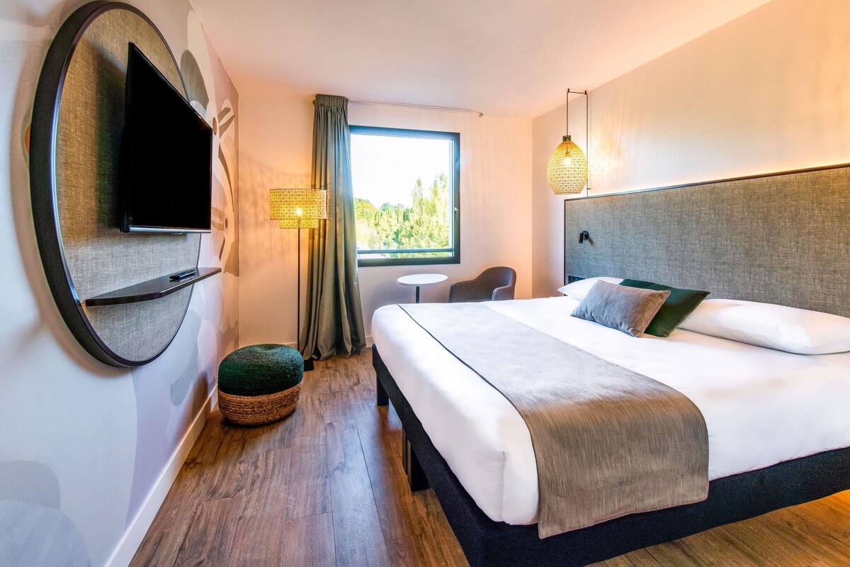 A hotel room with a bed, a TV, a lamp, a window, and a small table.
