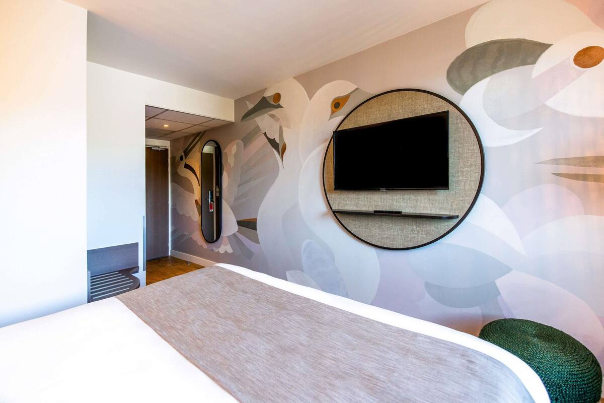 A modern hotel room with a large bed, a round mirror, a flat-screen TV mounted on the wall, and a decorative wall mural.