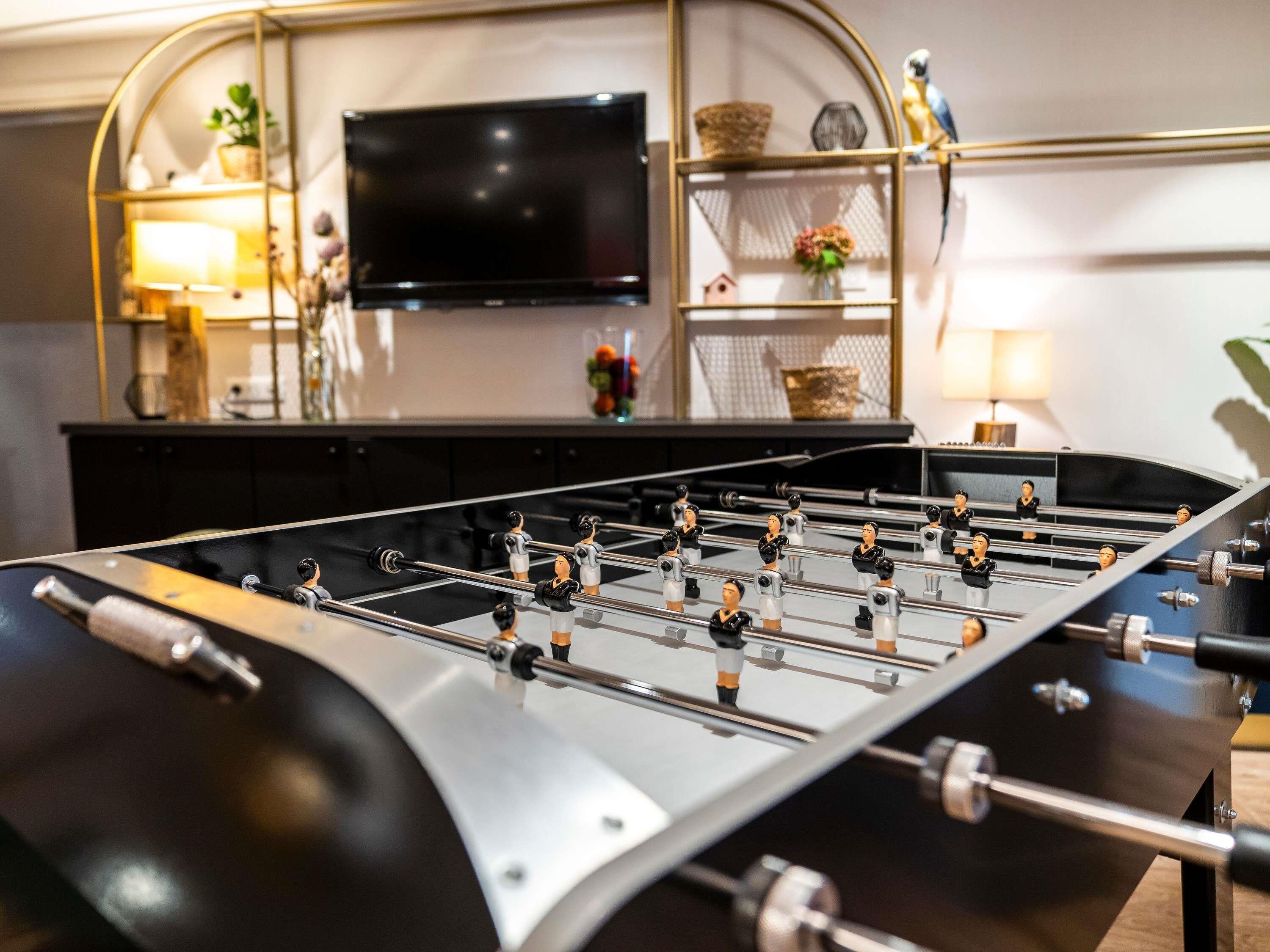 A foosball table in a room with a television, shelves with decorative items, and a lamp.