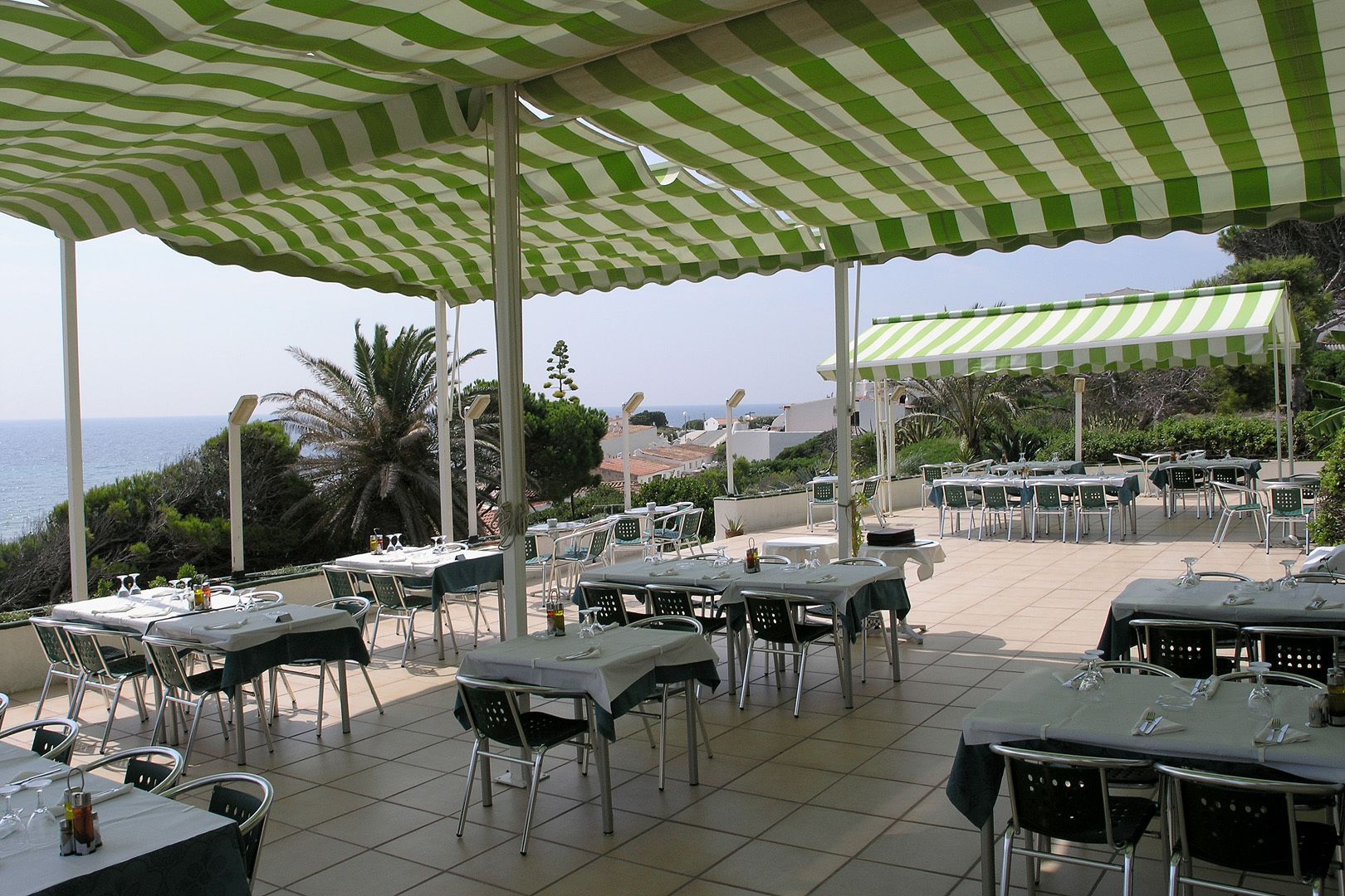 An outdoor dining area with tables and chairs under striped canopies, overlooking a coastal view.