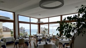 Een restaurant met meerdere tafels en stoelen, grote ramen met uitzicht op zee, en een plafond met ronde verlichtingen.