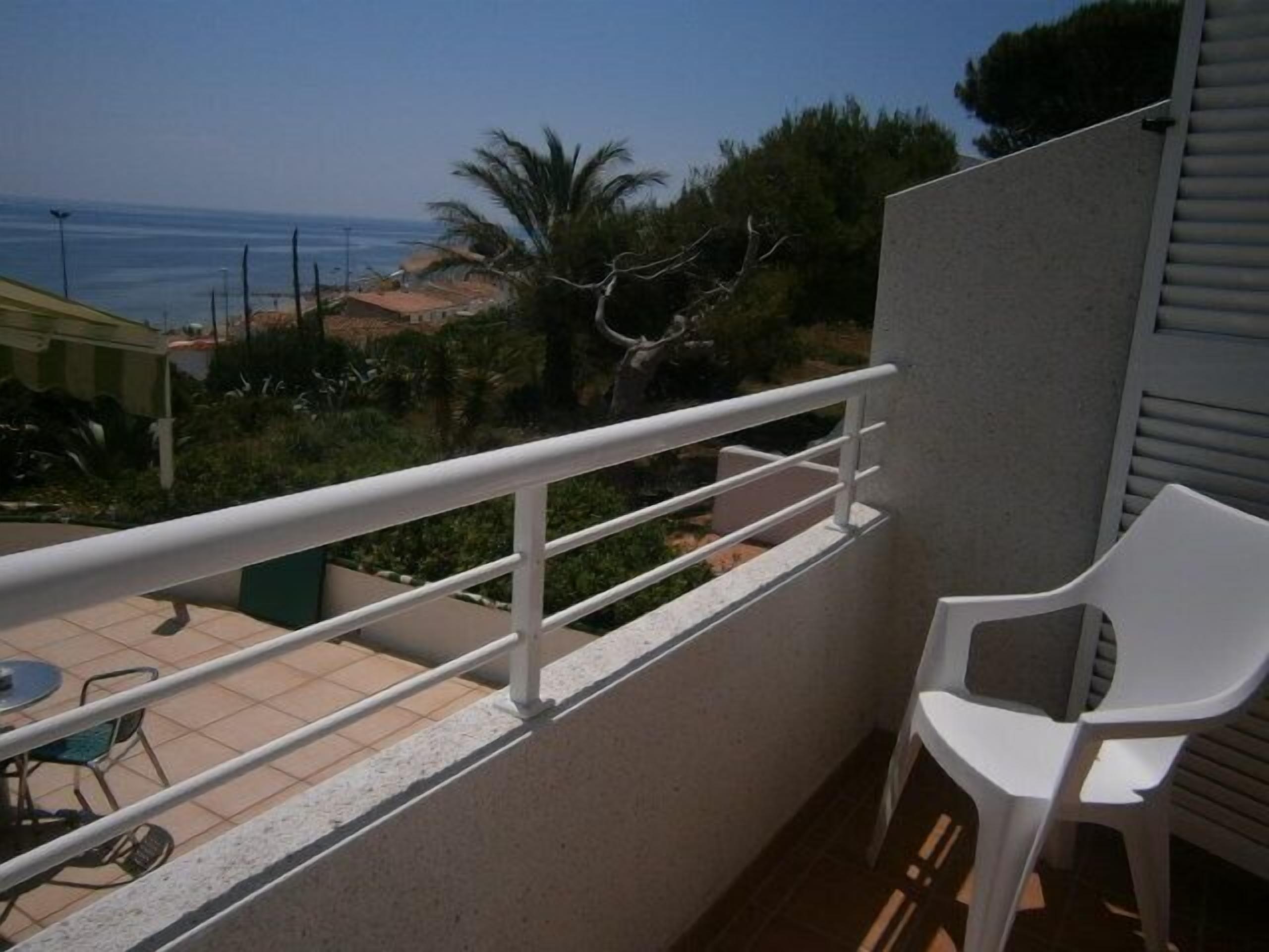 Un balcón con barandilla blanca, una silla de plástico blanca y vistas al mar y al follaje.