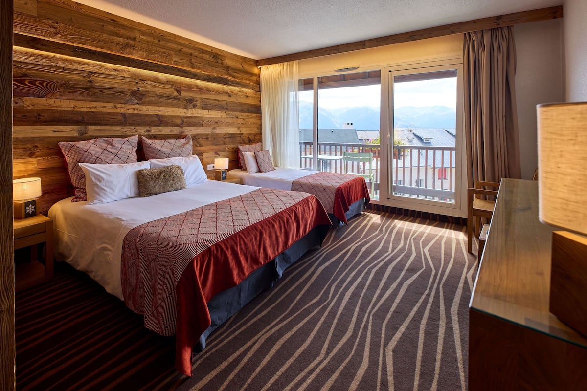 Habitación de hotel con dos camas, cabecero de madera, ventanas amplias con cortinas y vistas a las montañas.