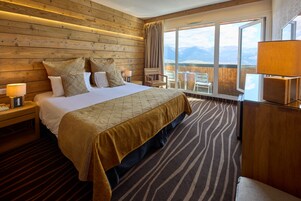 Quarto de hotel com cama grande, cabeceira de madeira e vista para montanhas através de uma janela ampla.