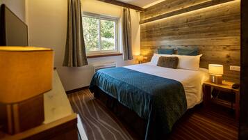 Habitación de hotel con una cama grande, dos mesitas de noche con lámparas, una ventana con cortinas y un cabecero de madera.