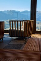 Quarto com uma janela ampla que oferece vista para montanhas. Há duas cadeiras de madeira com almofadas.