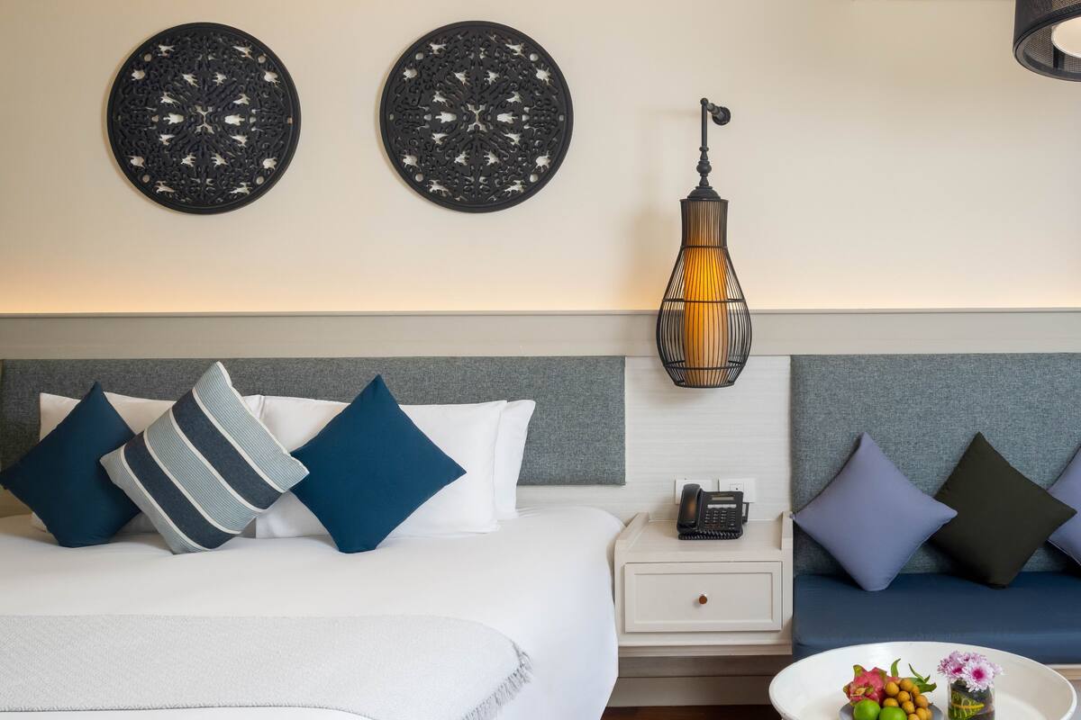 Ein Hotelzimmer mit Bett, Nachttisch, Lampe und einem kleinen Tisch mit Obst und Blumen.