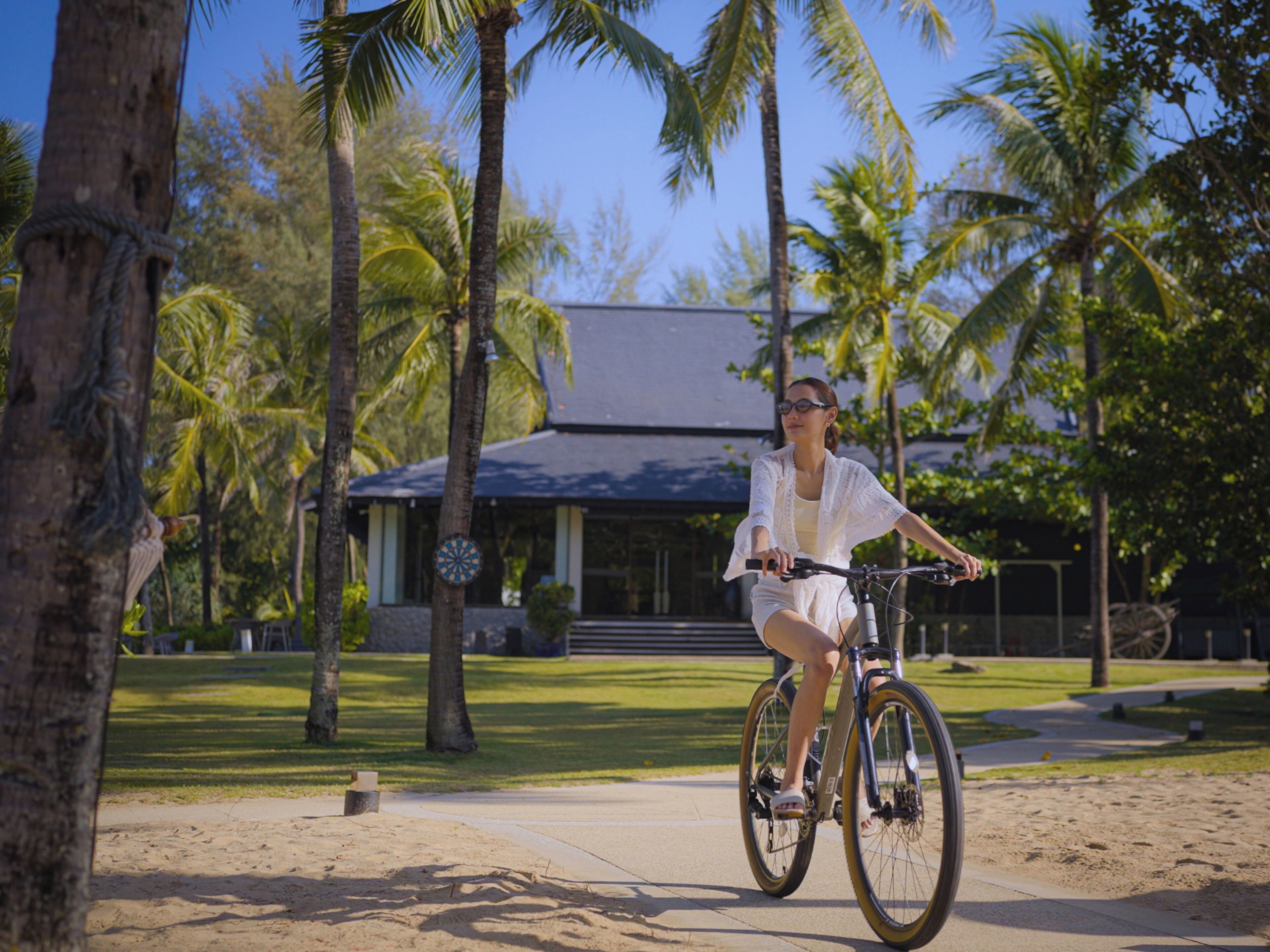 A person riding a bicycle on a sandy path with palm trees and a building in the background.