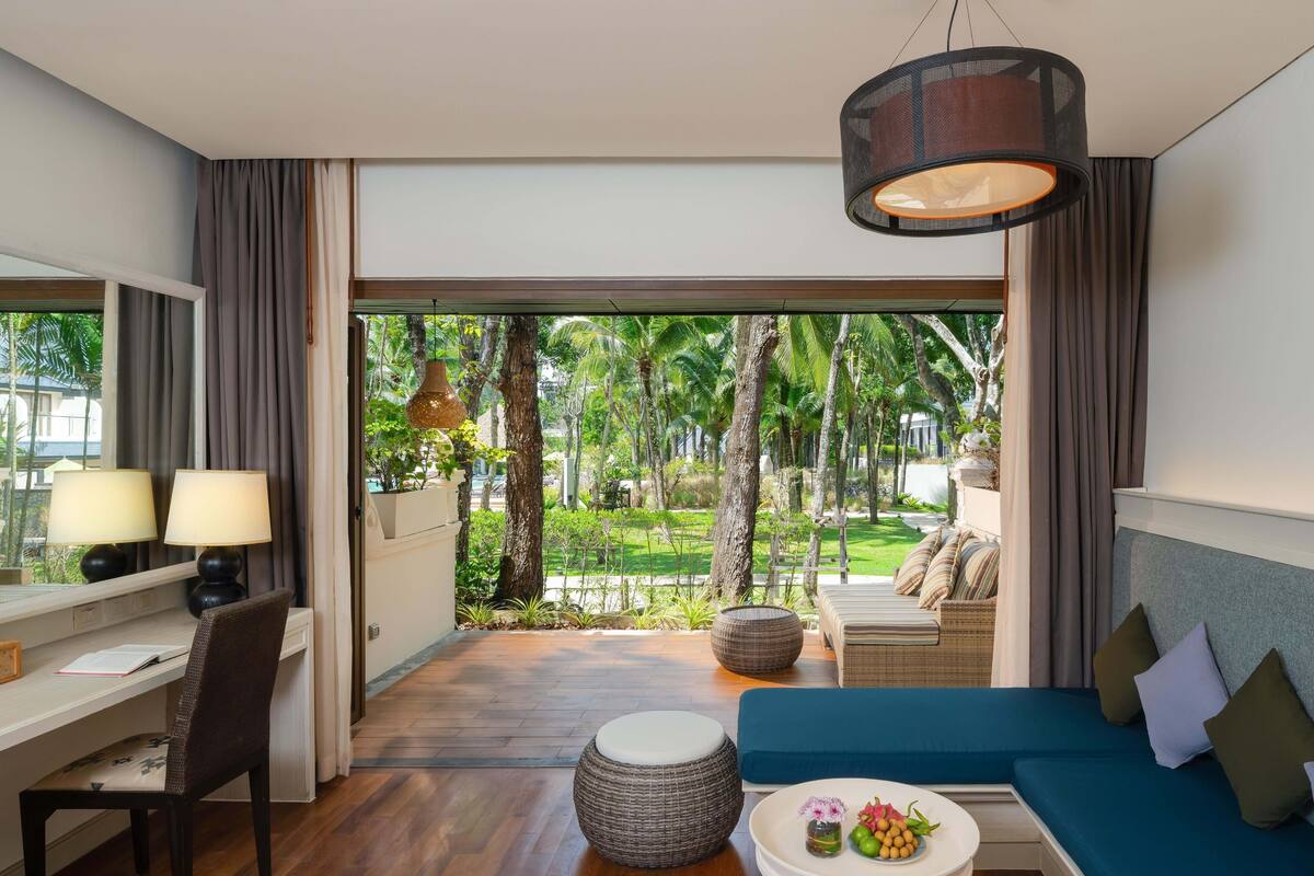 Ein modernes Hotelzimmer mit Schreibtisch, Stuhl und einer Couch. Blick auf einen tropischen Garten.