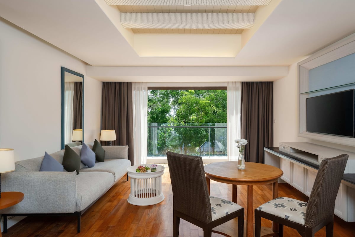 Ein modernes Hotelzimmer mit einer Couch, zwei Sessel, einem kleinen Tisch, einem Fernseher und einem Balkon mit Blick auf Bäume.