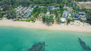 Vue aérienne d’une station balnéaire comprenant plusieurs bâtiments de style bungalow, une plage de sable et une mer turquoise aux eaux limpides.