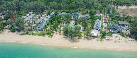 An aerial view of a coastal resort with multiple bungalow-style buildings, a sandy beach, and a clear turquoise sea.