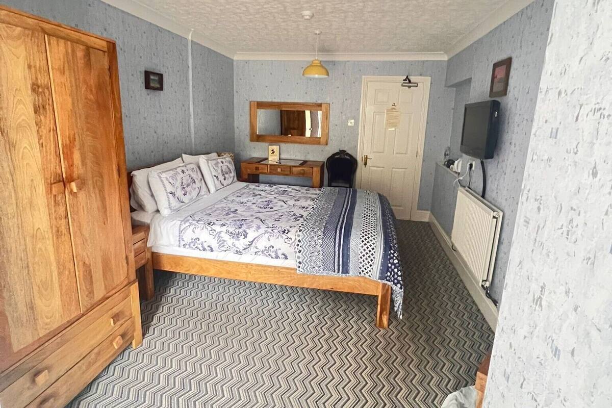 A bedroom with a bed, wooden wardrobe, dresser, mirror, and a television on the wall.