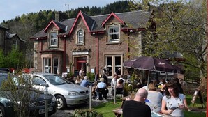 A building with a traditional architectural style, featuring a brick facade and a red roof. There are outdoor seating areas with tables and umbrellas, and a car parked in front. - The Old Rectory Inn (Callander)