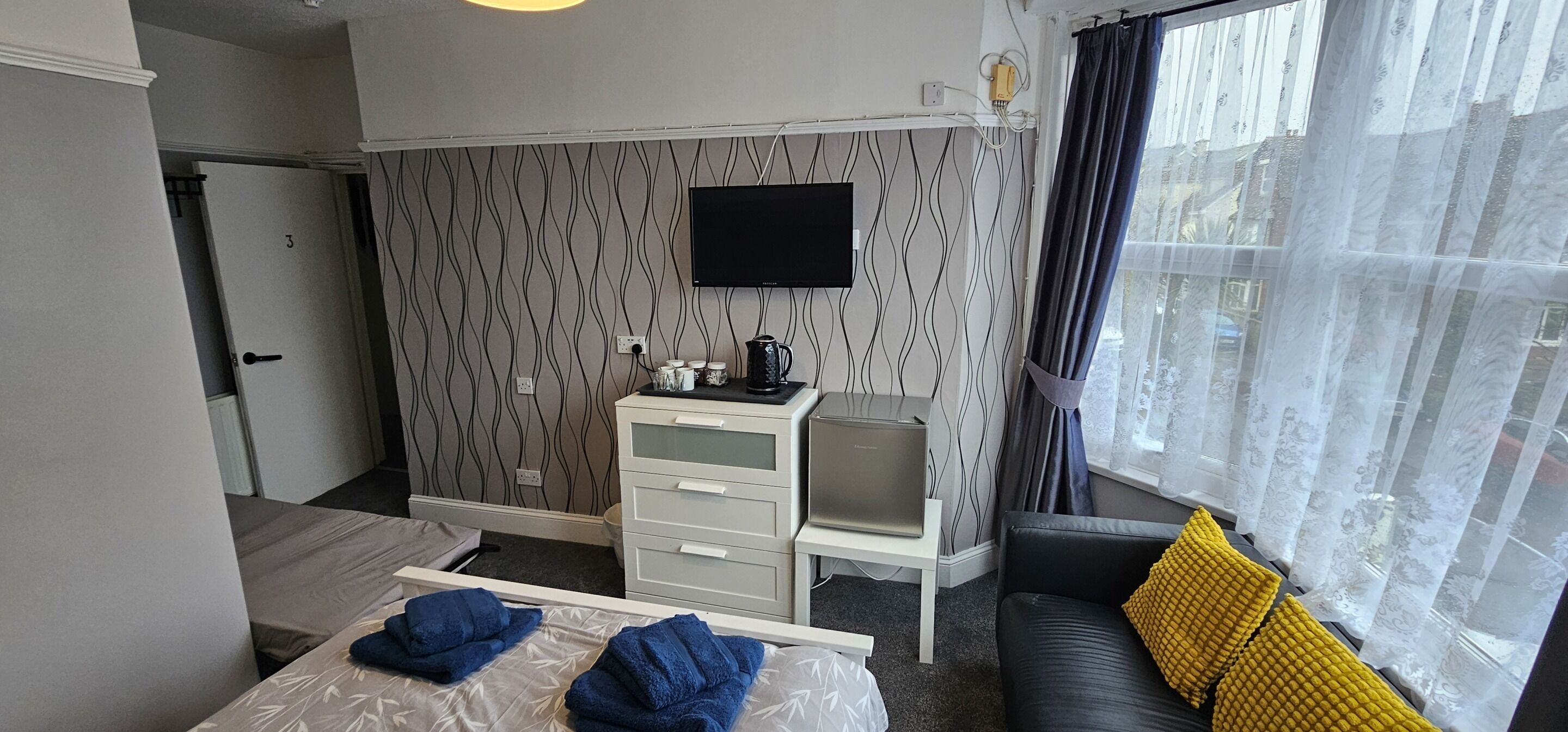 A hotel room with a bed, a desk with a laptop, a TV on the wall, and a window with curtains.
