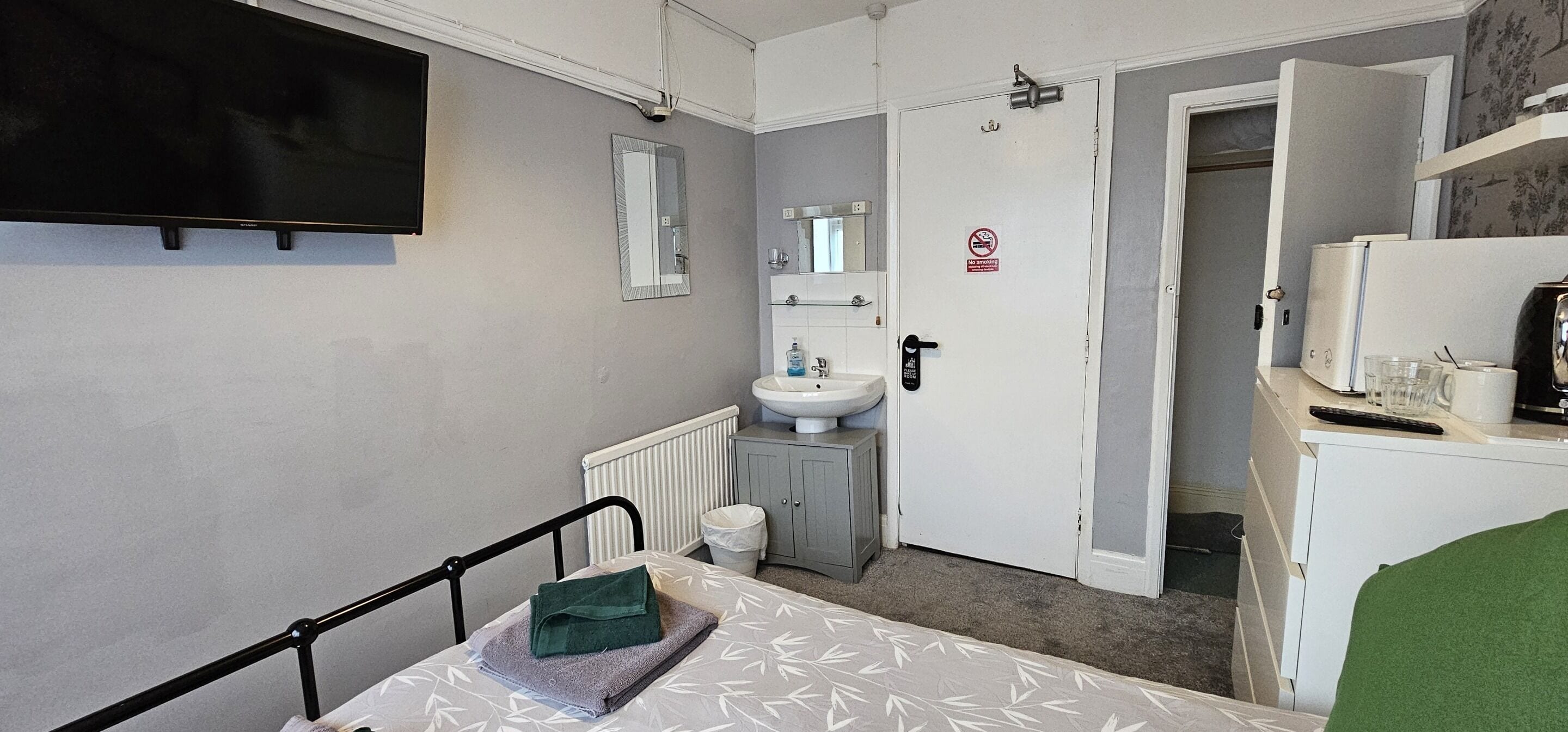 A small, compact room with a bed, a bedside table, a television, a sink, and a mirror.
