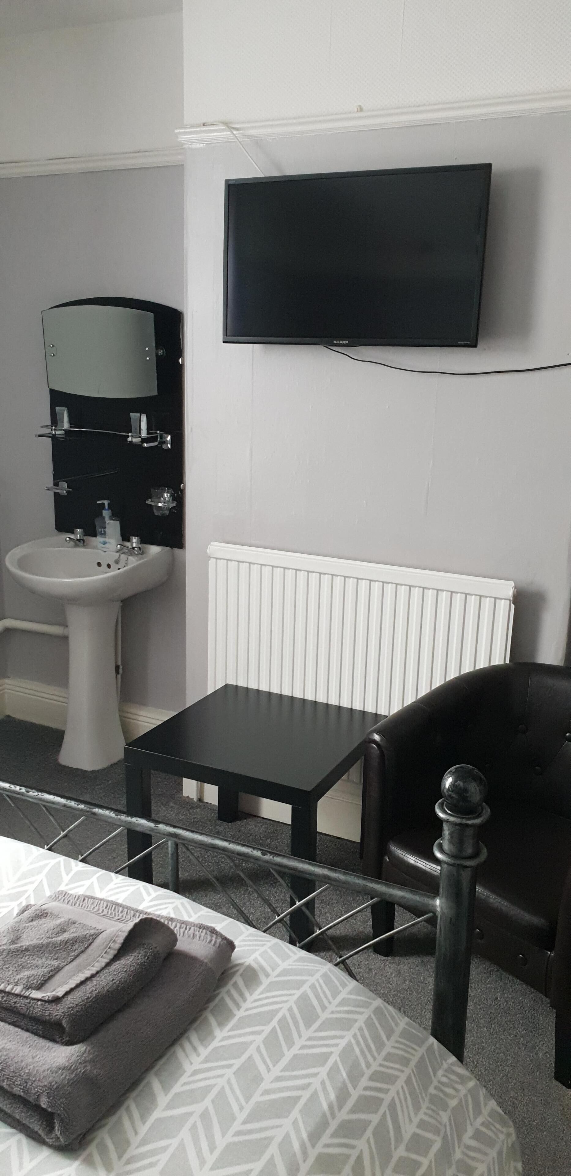 A hotel room with a bed, a bedside table, a black chair, a TV mounted on the wall, a white radiator, a sink, and a mirror.