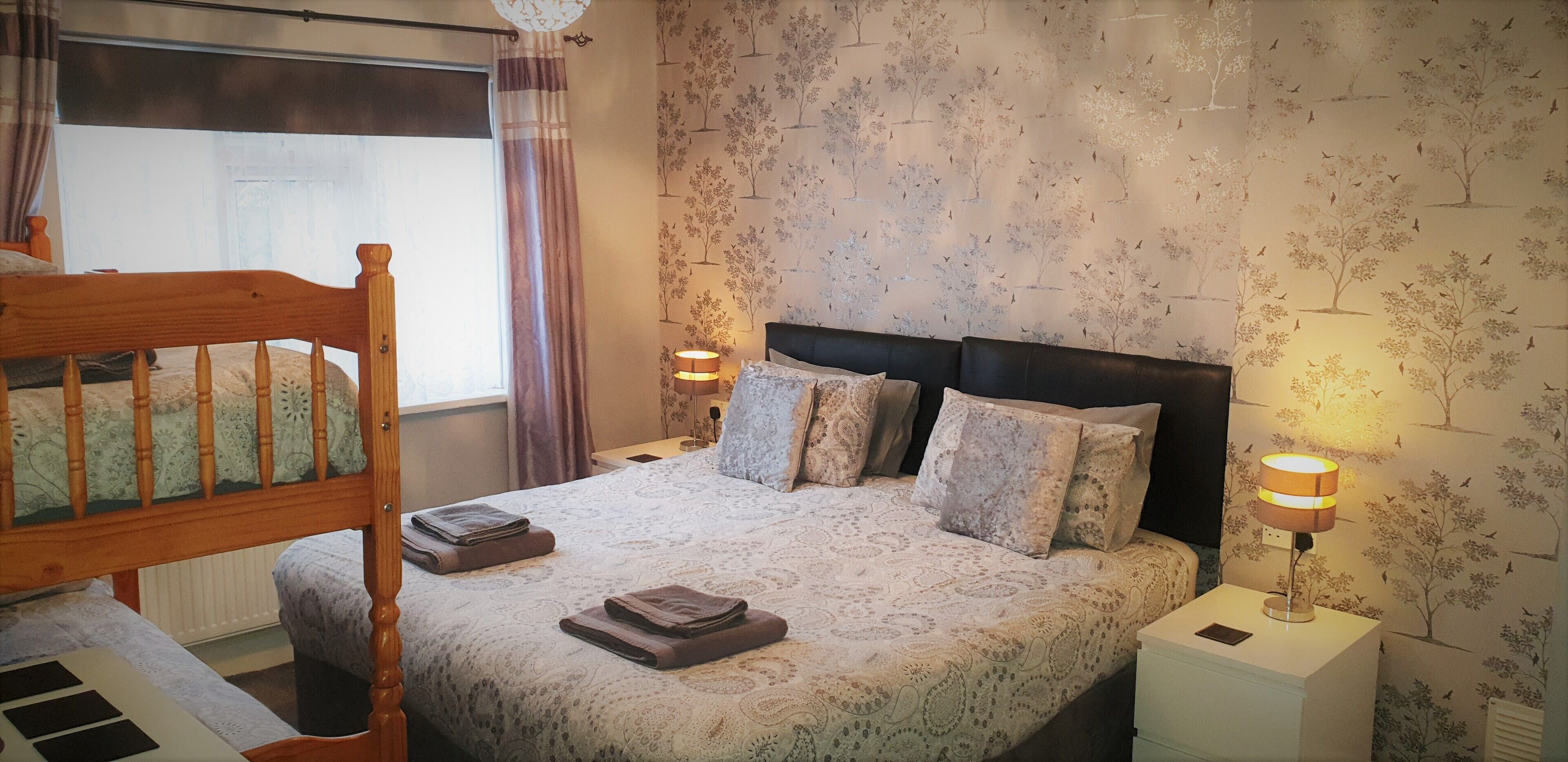 A bedroom with a wooden headboard, a patterned bedspread, multiple pillows, two bedside lamps, and a window with curtains.