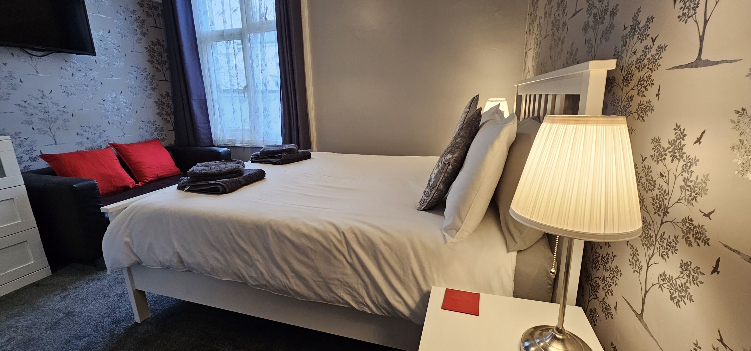 A neatly made bed with pillows and a folded towel, a bedside table with a lamp, a dresser, and a window with curtains.