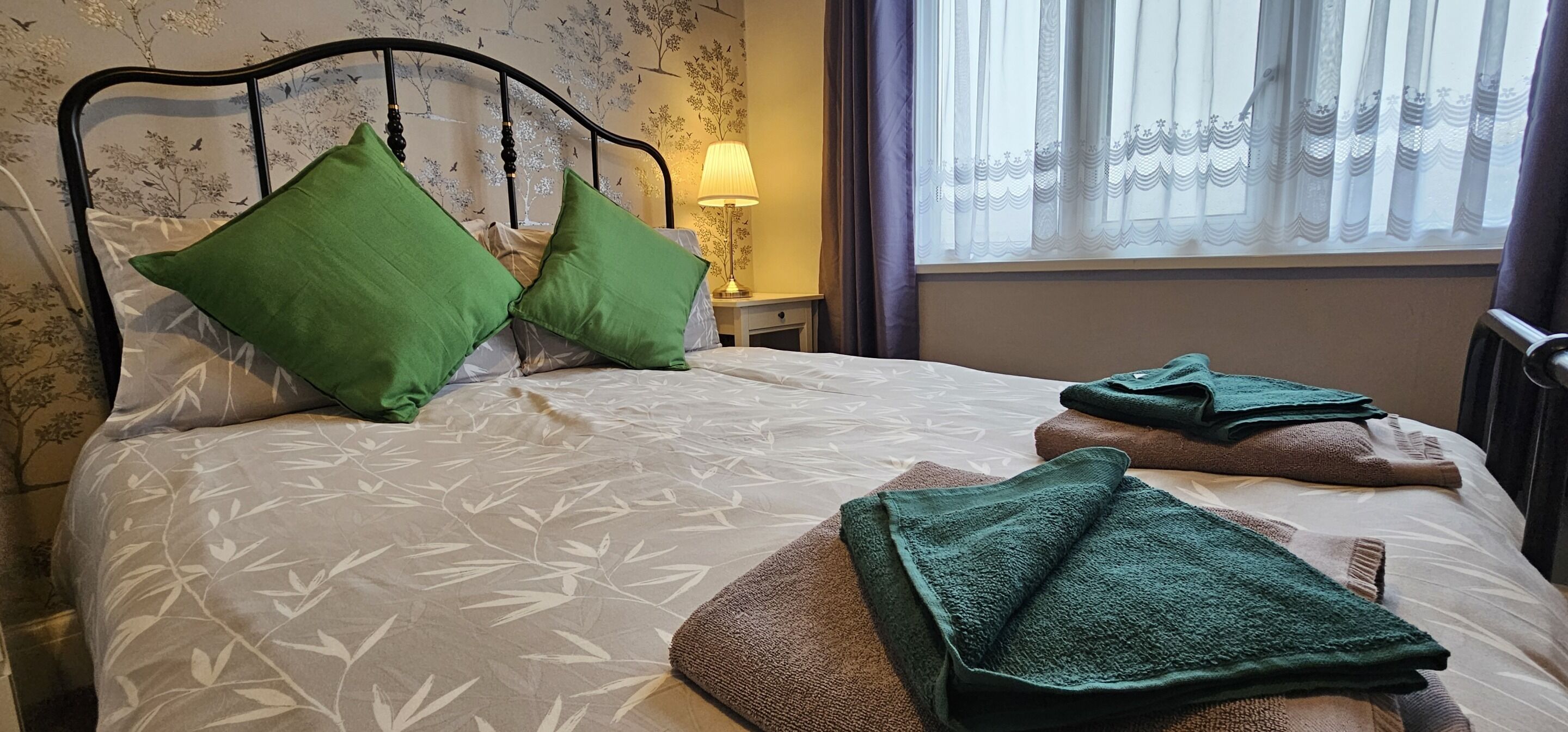 A neatly made bed with green pillows, a patterned bedspread, and folded towels on the side.