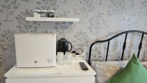 Ein kleiner weißer Tisch mit einem weißen Kühlschrank, einer schwarzen Kaffeemaschine, einer Fernbedienung und einem Regal mit Gläsern.