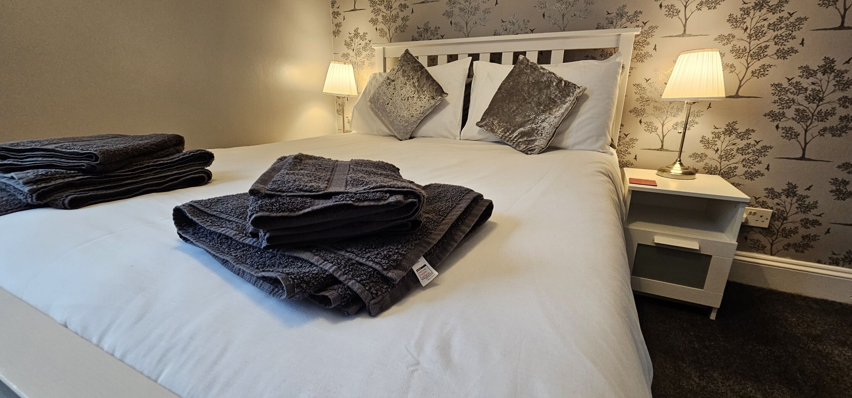 A neatly made bed with folded towels, two bedside lamps, and a nightstand with a lamp.