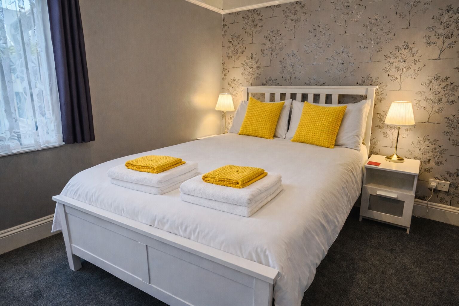 Una cama bien tendida con ropa blanca y dos toallas amarillas, una mesita de noche con una lámpara, y un estampado floral en el papel tapiz de las paredes.