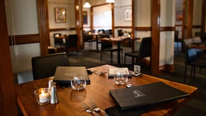 Une table de restaurant est dressée avec une bougie, des verres, des couverts et un menu. Le restaurant est décoré avec des meubles en bois et des luminaires suspendus.