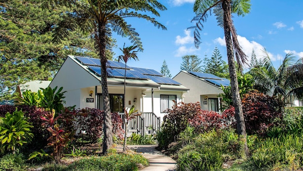 Une maison à deux étages, dotée de panneaux solaires sur le toit, entourée d’une végétation luxuriante et d’arbres.